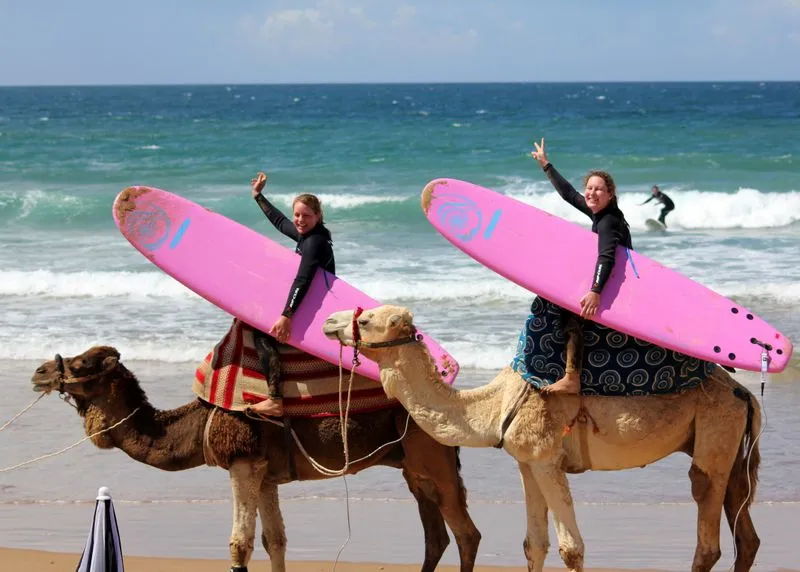 Zwei Surferinnen mit pinken Surfboards reiten auf Kamelen am Strand